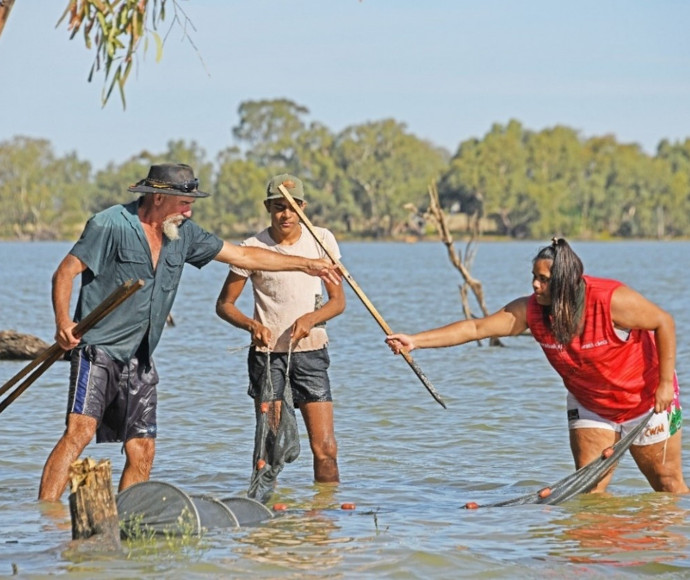 Three people in the water up to their knees, one older with a beard, passing poles and nets to a young woman and man