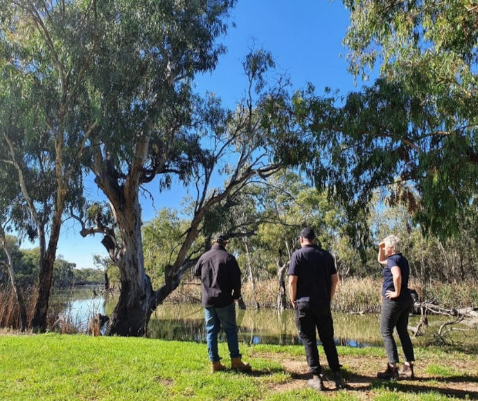 Three people standing by trees on the flat riverbank and looking at the water