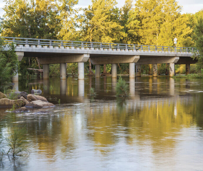 The long Yarramundi bridge sits on top of the Nepean river, amidst a backdrop of trees in various shades of green, reflected in the water.