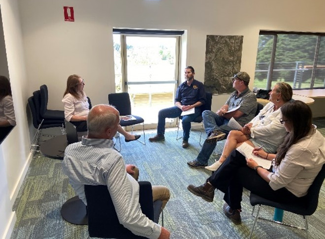 A circle of 6 people sitting in chairs inside a white-walled room with a glass door and a large glass window behind them at the Aboriginal joint management regional Aboriginal workshop in Katoomba.