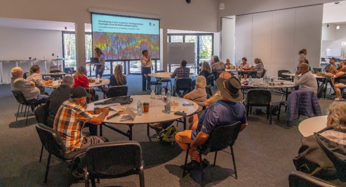 People seated at round tables in a conference room, facing a presenter with a large screen.