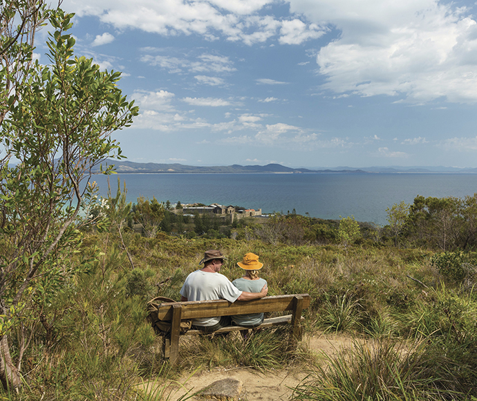 Two adults sit on a bench at the monument track, part of Arakoon National Park. A male figure places his arm around a female figure and they are wearing wide brimmed hats. Beautiful trees of different shades of green line the foreground and the background, which looks off into the serene blue water.