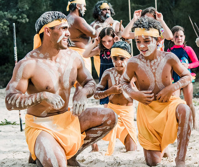 A group of people, including adults and children, are participating in a cultural or traditional event on a sandy beach. They are wearing yellow cloths around their waists and have white body paint in various patterns on their torsos and arms. Some participants are kneeling, while others stand in the background. The backdrop includes trees and vegetation.