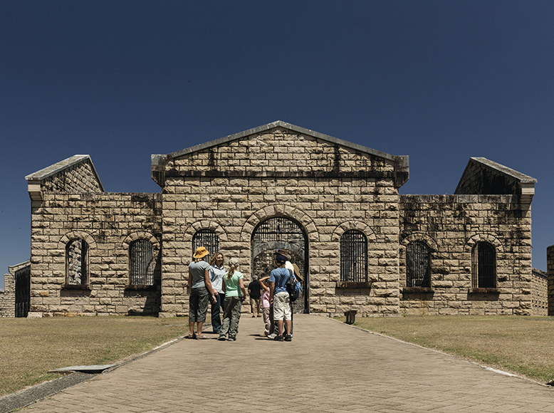 Visitors gather to explore Trial Bay Gaol in Arakoon National Park. This historic site stands in the centre, with its brick structure still in tact. It appears to be a sunny day as the visitors are wearing hats and are gathered around a tour guide.