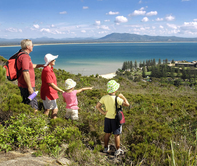 A group of four people, consisting of two adults and two children, are standing on a grassy hill overlooking a scenic coastal landscape. The adults are wearing hats and carrying backpacks, while the children are wearing sun hats. One of the children is pointing towards the ocean. The background features a beautiful view of the ocean with clear blue water, a sandy beach, and a distant mountain range under a partly cloudy sky. The scene is serene and picturesque, capturing a moment of outdoor exploration and 