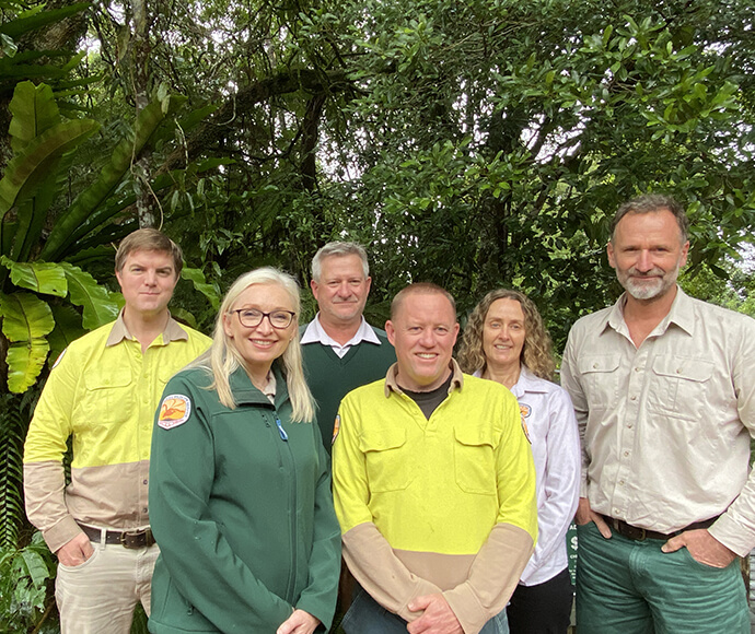 Group photo of the Dorrigo Escarpment Great Walk project team, dressed in unforms, stands in front of a lush, green forest, smiling at the camera.