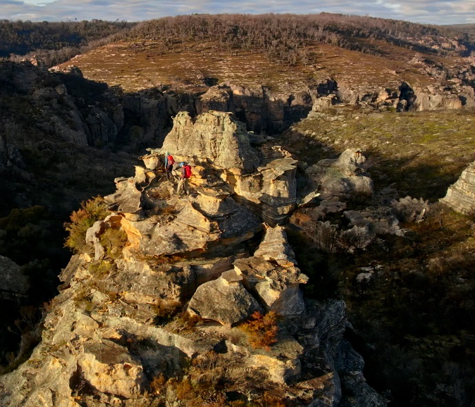 Panoramic view of the Gardens of Stone reserves, featuring rocky mountains of varying heights, grassy hills and shady valleys. There are two people hiking on one of the rocky hills carrying red backpacks and wearing hiking clothes. The sun illuminates some of the mountains, while casting shade across the valley.