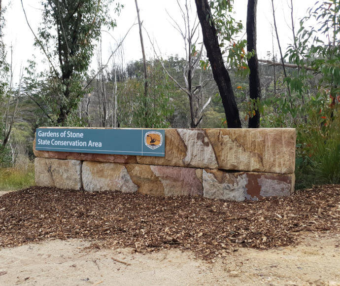 A sign announcing the 'Gardens of Stone State Conservation Area' on a low stone wall with the National Parks and Wildlife Service logo of a black lyrebird over an orange background, with tanbark in front of the sign and trees behind.