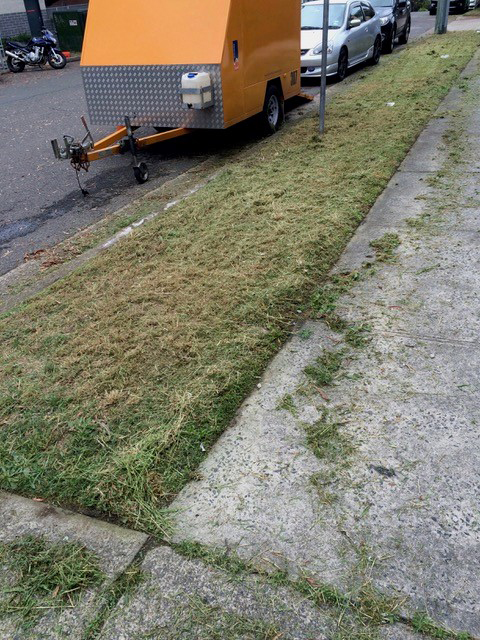 Grass verge about a meter wide that has been freshly mown. There is a concrete footpath and a bright orange trailer and parked cars on street.