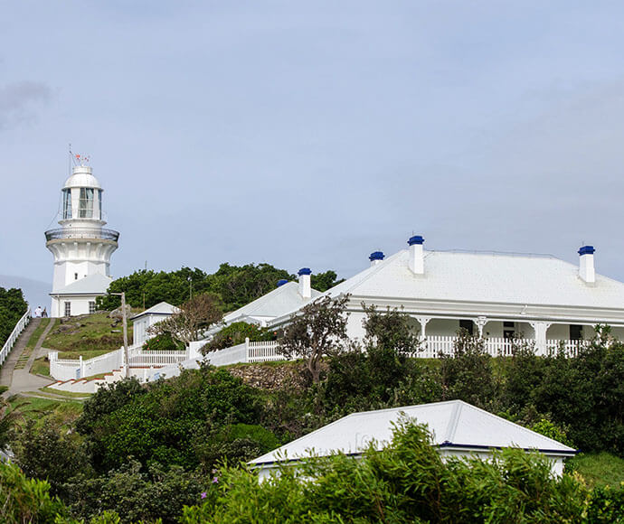 A white lighthouse with a cylindrical tower and a lantern room at the top is situated on a hill. The lighthouse is surrounded by white buildings with blue accents on their roofs and chimneys. The buildings are connected by a white picket fence and are surrounded by green vegetation. The sky is overcast.