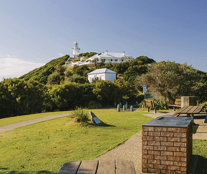 A scenic view of a lighthouse and its surrounding buildings on a hilltop, with a clear blue sky in the background. The foreground features a grassy area with a paved path, a brick barbecue grill, wooden benches, and a small information plaque. The lighthouse and buildings are white, contrasting with the green foliage around them. The image captures a peaceful and picturesque outdoor setting, likely a popular spot for visitors.