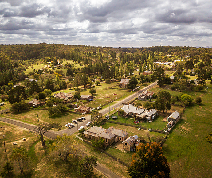 Aerial view of a historic rural village with several old stone and wooden buildings surrounded by grassy fields and scattered trees. The houses have rustic, weathered appearances, and a few cars are parked along the roads and in driveways. The main road runs through the center, dividing the village into two sections. In the background, a lush landscape of rolling hills and dense green forest stretches towards the horizon under a partly cloudy sky. The scene captures a tranquil, preserved rural community wit