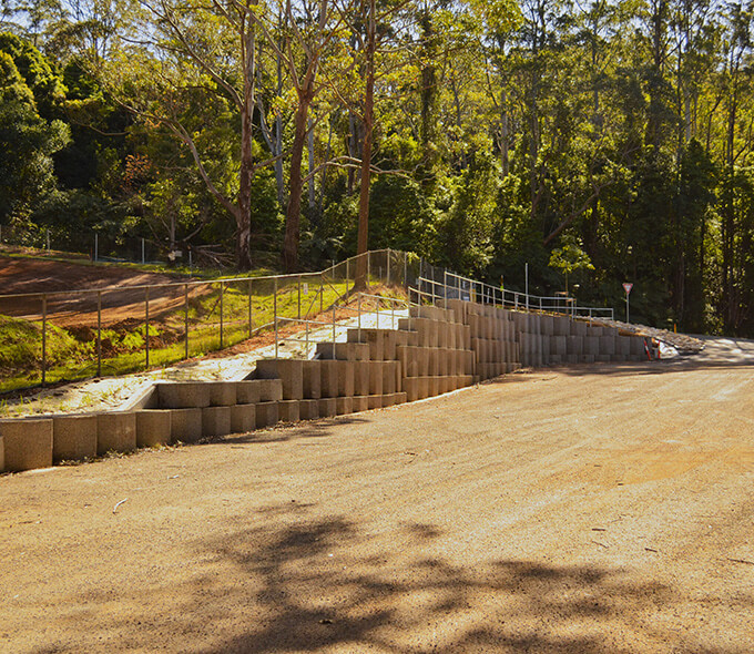 A dirt road runs alongside a terraced retaining wall made of concrete blocks, which supports a fenced-off slope. The area is surrounded by tall trees and lush greenery, blending construction with the natural environment. The road curves to the right in the background, highlighting efforts to manage erosion and stabilize the terrain.