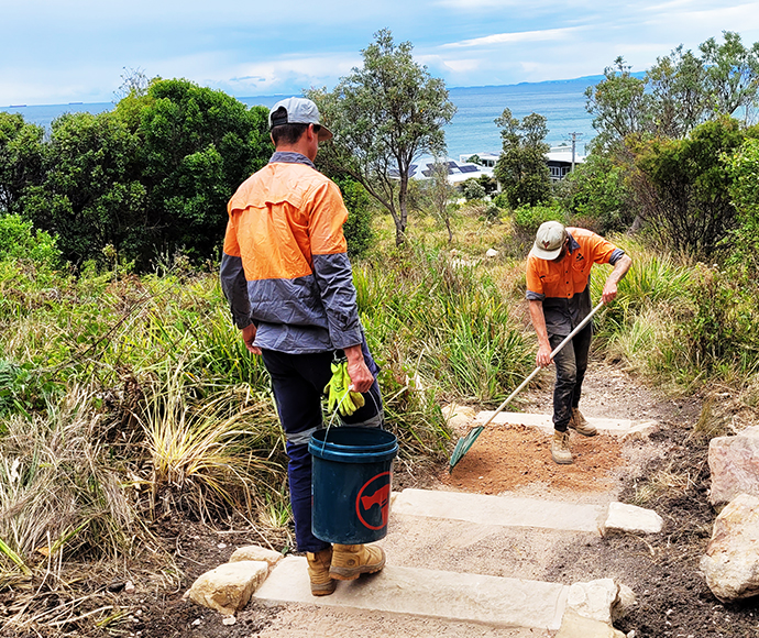 Two workers in orange and gray uniforms are maintaining a dirt path with stone steps. One worker is raking the path, while the other holds a bucket and some plants. The scene is set in a natural area with greenery and a view of the ocean in the background.