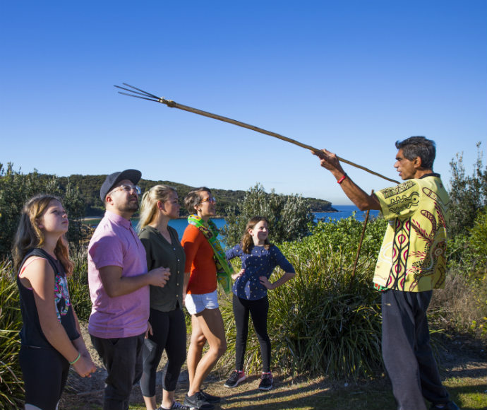 An Aboriginal guide leads visitors on a cultural tour at La Perouse, sharing stories of local Aboriginal history and connection to the coastal landscape. The tour provides insights into traditional knowledge and the deep relationship between people and Country.