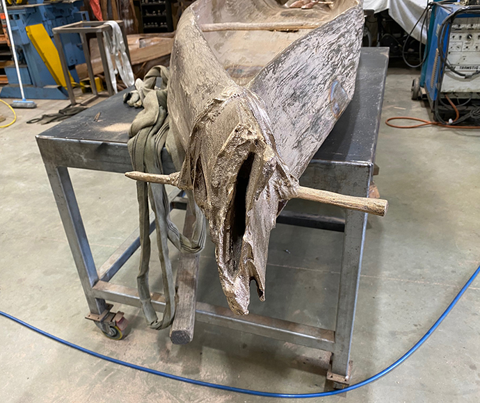 A detailed look into the fabrication of "The Canoes" sculptures. The near-complete canoe sculpture sits in the centre of the image in a workshop on a large metal table with castors, while various ropes and cords surround the workshop along with a concrete floor and some machinery in the background.