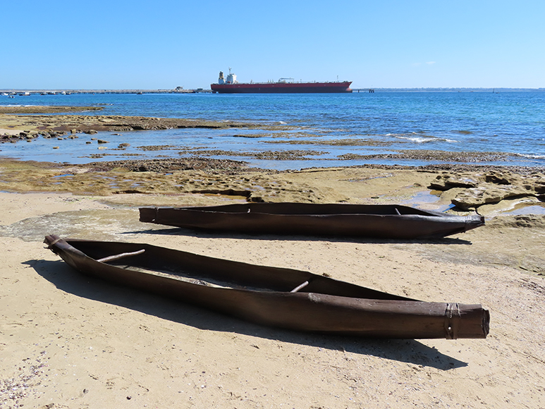 Two sculptures of canoes sit in the foreground, which are exceptionally long and ornate. They rest on the sand with the gentle ocean shore in the background, featuring a cargo ship in the distance, showing the contrast between the natural and man-made world. The crystal clear blue sky enhances the scene, and shows the beauty of nature.