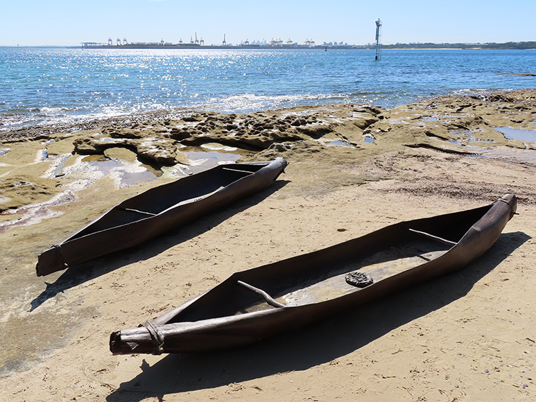 Two sculptures of canoes sit parallel to each other in the foreground on a diagonal angle. They rest on the light brown sand with the gentle ocean shore in the background. The sun shines on the glistening sea, and highlights the natural rock pools the sculptures are surrounded by in the distance.