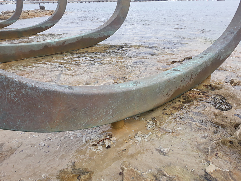 An intimate look at the stunningly beautiful sculpture titles "the eyes of the land and the sea." The sculpture features a series of gently curved beams which rest on top of a natural rocky surface, with a gentle layer of clear water dotting the surface and creating a shining effect between the sculpture and the surface it rests on.