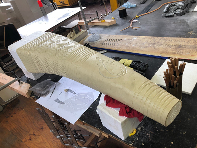 A glimpse into the fabrication of "the eyes of the land and sea" sculpture. One of the partially completed "ribs" of the sculpture sits on a workshop table in the centre, while various sculpting instruments are scattered across the table. The background features additional wood-working tools and machinery, while some natural light shines into the workshop.