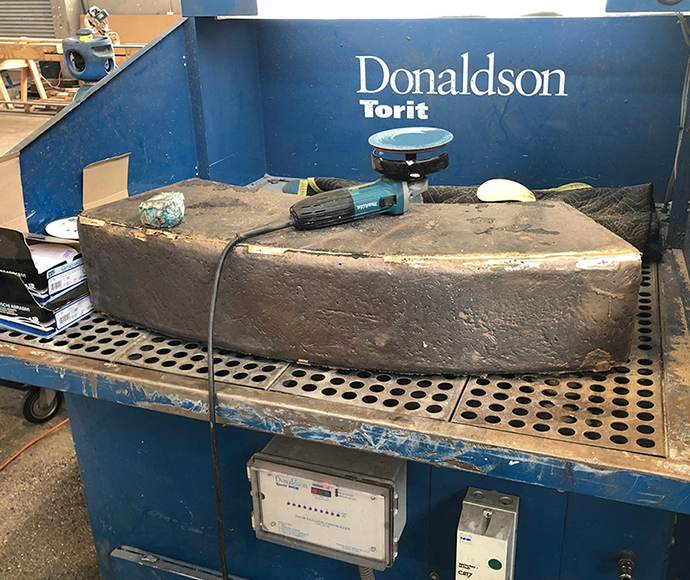 A unique look into the fabrication of one of the key elements of a sculpture. A piece of bronze sits on a large piece of machinery in a workshop, with a sanding tool resting on the top of the sculpture piece. Some buttons and switches are towards the bottom of the machine, with the machine featuring the words 'Donaldson Torit' on the top. The setting appears to be in a workshop, with additional tools and industrial accents composing the background.