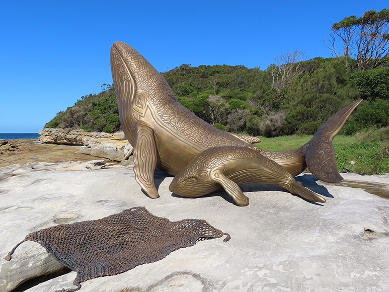 Detailed bronze sculptures of two whales, one bigger and one smaller, and a weaved fishing net sit amongst a beachy backdrop. The backdrop features a smooth rocky surface which the sculptures sit on, along with vibrant green grass and bushland in the background. The scene is set by a crystal clear blue sky creating a tranquil vista to highlight the beauty of the bronze sculptures.