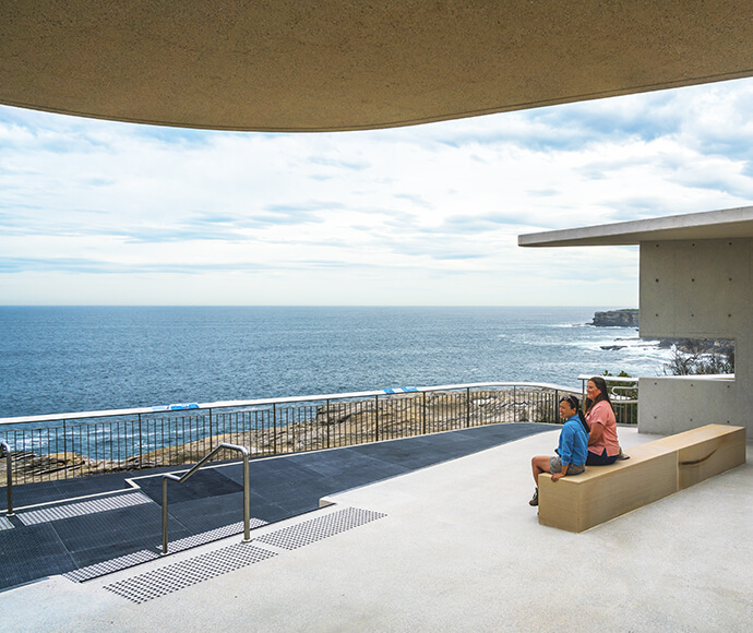 Whale watching platform at Cape Solander in Kamay Botany Bay National Park. The image shows a modern viewing platform with a curved roof, overlooking the ocean. Two people are seated on a wooden bench, enjoying the expansive view of the sea and distant cliffs. The platform has railings and a non-slip surface, ensuring safety for visitors.