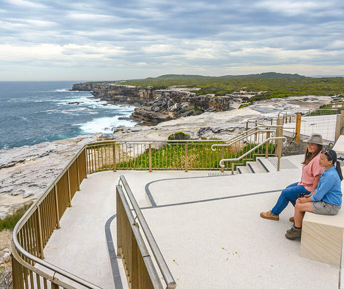 The image shows a scenic coastal view from a whale-watching platform. The platform is situated on a cliffside, overlooking the ocean with waves crashing against the rocky shore. The platform has railings and seating areas, and two people are sitting on one of the benches, enjoying the view. The sky is partly cloudy, and the landscape includes both rocky and grassy areas. The image captures the natural beauty and serene atmosphere of Cape Solander, making it an ideal spot for whale watching.