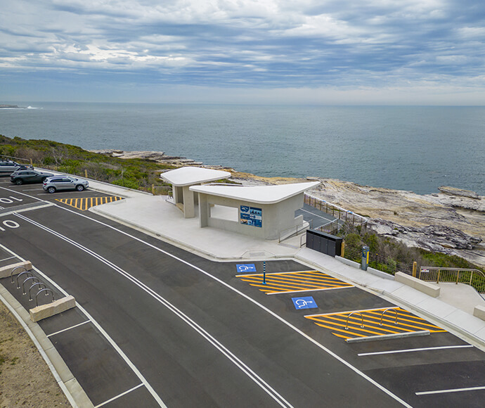 Whale watching platform at Cape Solander in Kamay Botany Bay National Park. Located on the edge of a coastal cliff, the platform offers an expansive view of the ocean. The modern design includes informational signage and a flat roof. Nearby, a parking lot with designated accessible parking spaces can be seen. The scenic location, complete with rocky coastline and partly cloudy sky, is ideal for observing marine life.