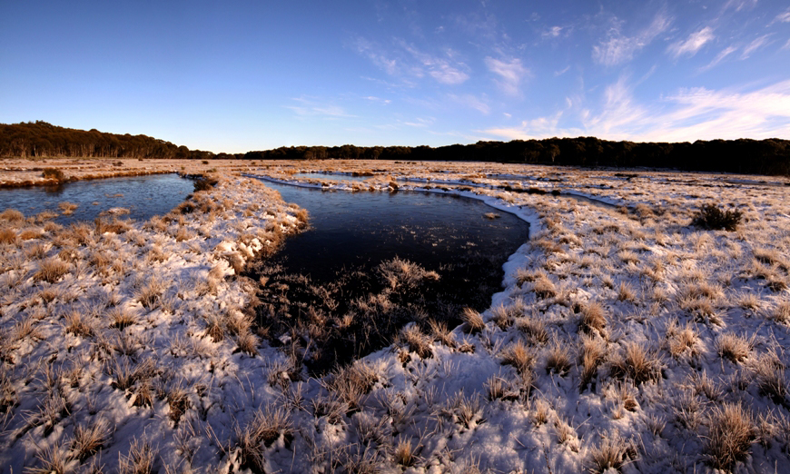 A picturesque early morning at Bullocks Hill bog, featuring a landscape with patches of frost and ice on the water, alongside areas of melted snow, all under a clear blue sky with flowing clouds, showcasing the diverse beauty of Kosciuszko National Park.