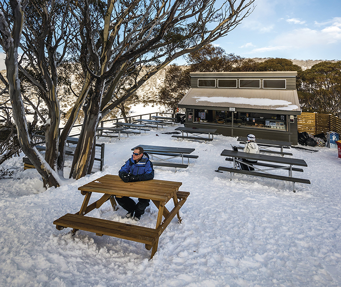 A tourist enjoying a winter day at the Alpine Eye cafe in Perisher Valley, seated at an outdoor table surrounded by snow-covered scenery, with a clear blue sky above, capturing the cozy atmosphere of the alpine environment.