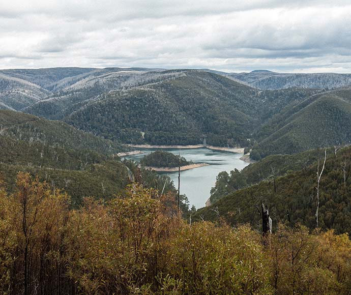 View from the top of a mountain overlooking a lake nestled between tall peaks along the Alpine Way in Kosciuszko National Park, showcasing water management efforts within the stunning natural landscape.