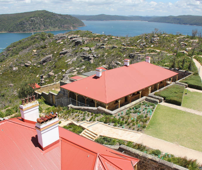 Ku-ring-gai Chase National Park Cottages: A cluster of red-roofed cottages atop a hill in Ku-ring-gai Chase National Park, overlooking vast bushland and water views. The image highlights the greenery and rugged terrain around the cottages.