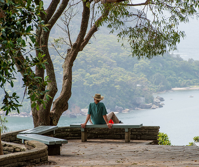 View from West Head Lookout in Ku-ring-gai Chase National Park, featuring a man wearing a hat sitting on a bench, overlooking the stunning coastal scenery with lush green hills and the sparkling waters of Pittwater.