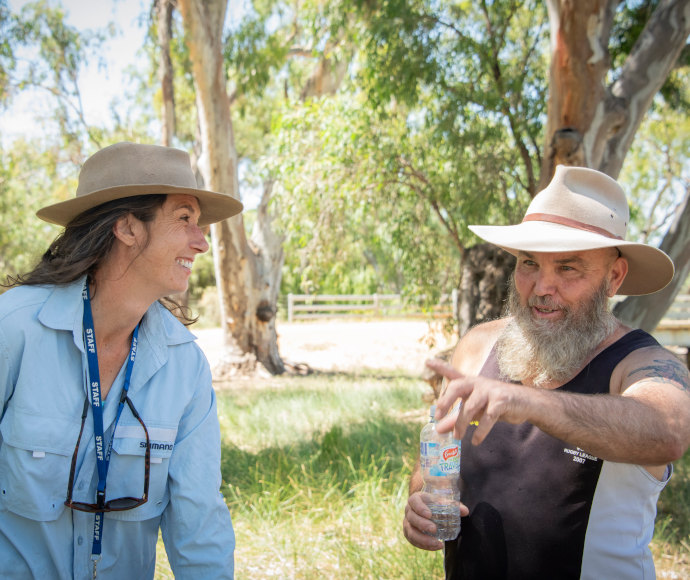 Jo Lenehan is on the left and wearing a blue work shirt and a bush hat. She is smiling at Uncle Ray Woods who is holding a water bottle and gesturing. He is wearing a single and bush hat. There are gum trees in the background.