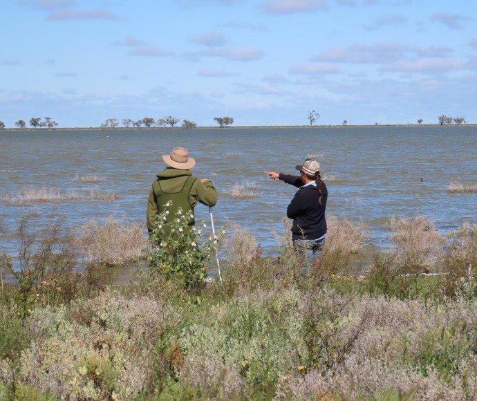 Two people looking out at the lake with their backs to the camera. Nathan is on the left and wearing a khaki-coloured coat and bush hat. He is holding a walking pole. Caroline is on the right and is pointing at something on the lake. She is wearing a black long-sleeved top, jeans and a cap.