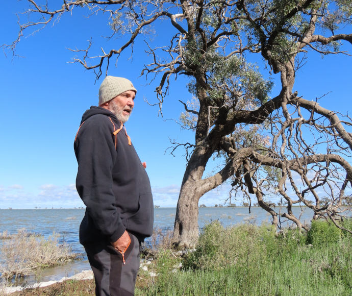 Uncle Ray Wood, standing next to a gnarled black-box tree on the shore of Dry Lake. He is wearing a black hoodie and a beanie.