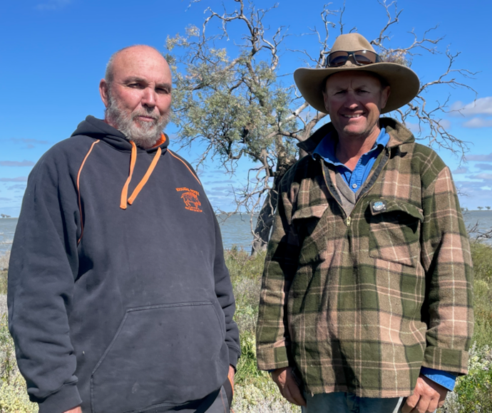 Uncle Ray Woods and Simon Booth are facing the camera with Dry Lake in the background. Uncle Ray is on the left and is wearing a black hoodie. Simon is on the right and is wearing a checked work jacket and bush hat.