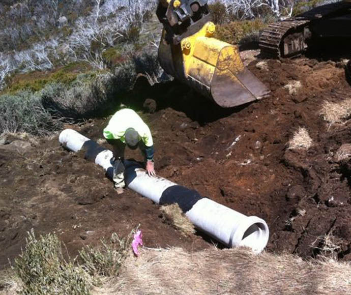 A worker laying a concrete pipe designed for animal crossing, with an excavator visible in the top right corner, set against a natural landscape in Kosciuszko National Park.