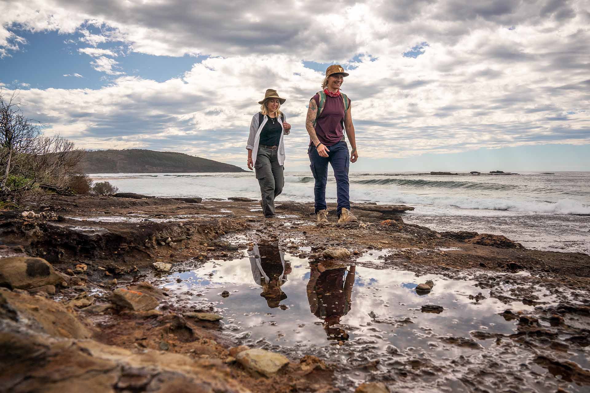 Two women dressed in hiking clothes, sun hats and boots walking along the rocks by a beach