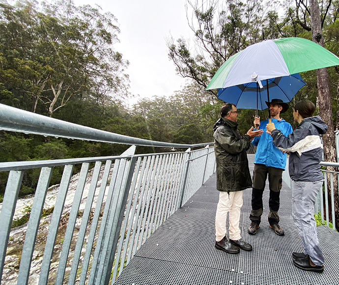 Three people with an umbrella on a forest walkway near a river.