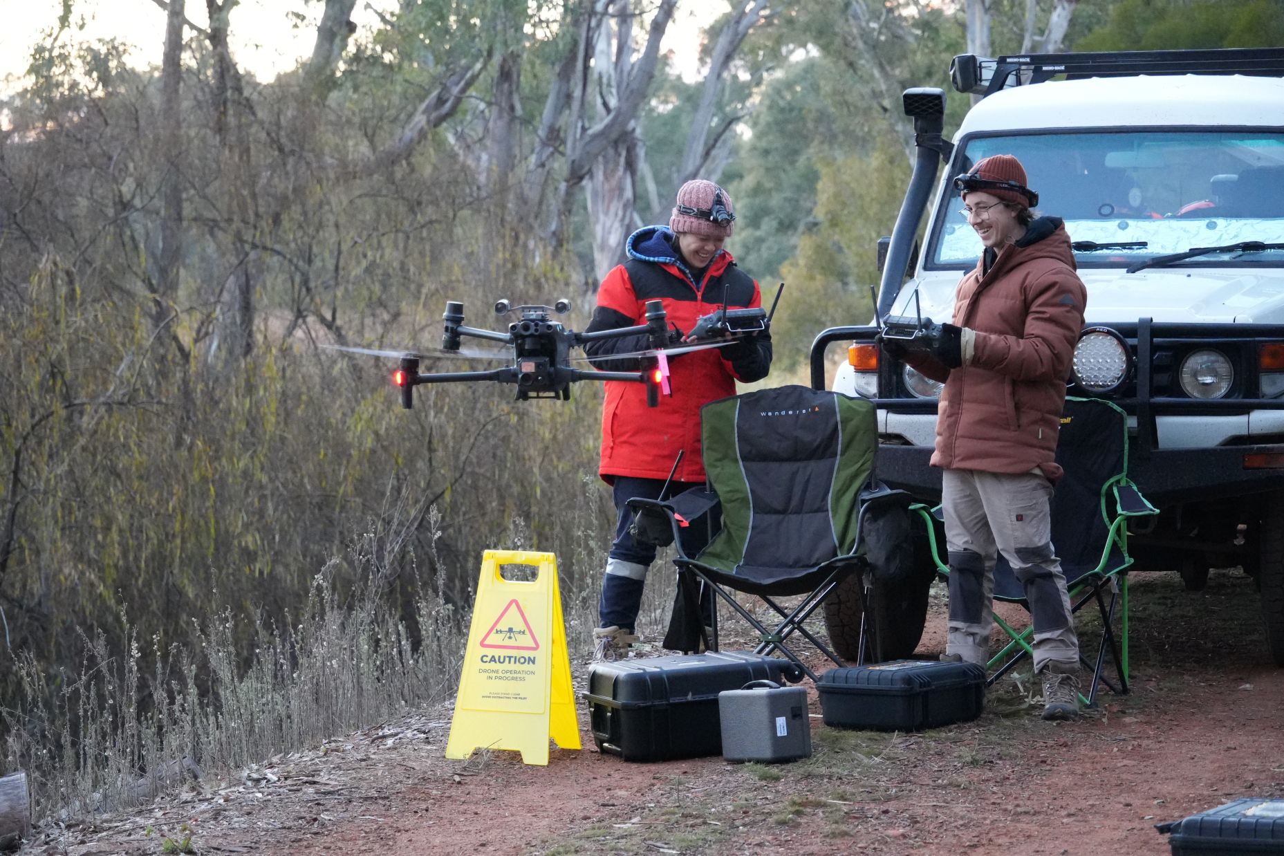 Two researchers stand in front of a vehicle testing a drone. The drone hovers in front of the 2 researchers who are holding remotes