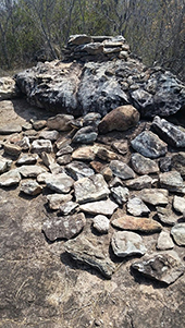 Stone cairn and scattered rocks on a rocky ground, surrounded by sparse bushes. The scene conveys a natural, rugged outdoor setting.