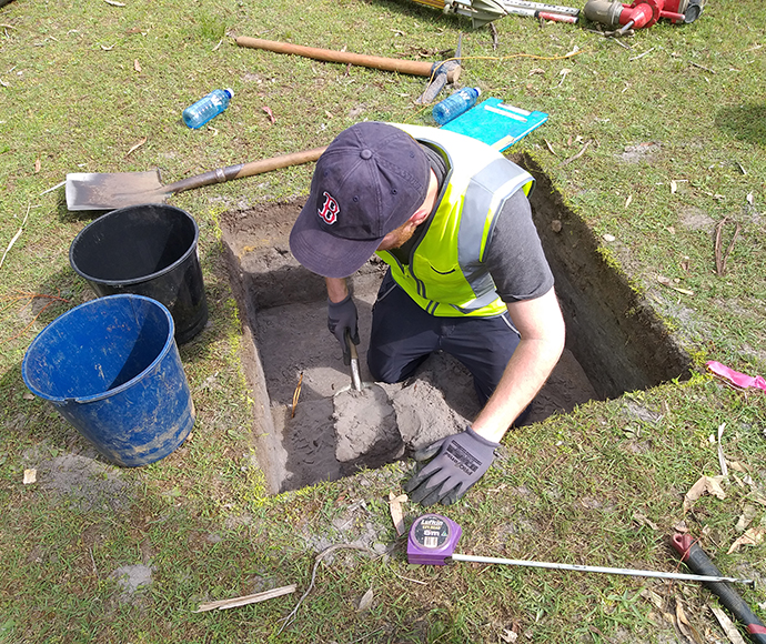 The image depicts an Aboriginal archaeological investigation at Bonnie Vale in the Royal National Park. It shows a single person wearing a high-visibility vest, actively working at an excavation site. The area is marked with strings and pegs to create grids for excavation, and various tools and equipment are visible around the site.