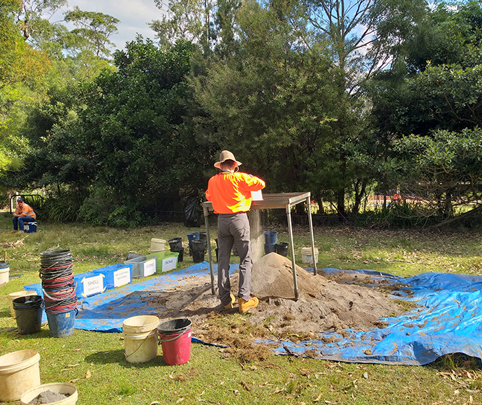 The image shows an Aboriginal archaeological investigation at Bonnie Vale in the Royal National Park. The scene features several people wearing high-visibility vests and hats, engaged in various activities around an excavation site. They are surrounded by tools and equipment, and the area is marked with flags and tape. The setting includes lush vegetation and trees, creating a natural backdrop for the investigation.