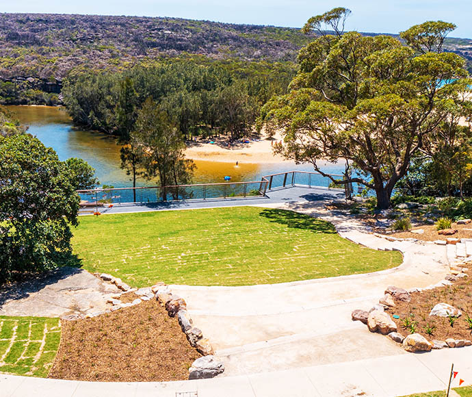 A modern viewing platform with glass railings at Wattamolla in Royal National Park, overlooking a lagoon bordered by a sandy beach and lush vegetation. The foreground features a well-maintained grassy area with a pathway and landscaping with rocks and plants. In the background, forested hills enhance the natural beauty of the scene.