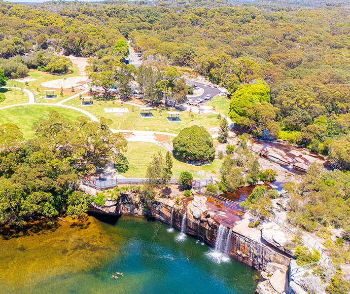 An aerial view of the Wattamolla picnic area in Royal National Park, featuring a lush green park with several picnic shelters, pathways, and parking spaces. The park is surrounded by dense forest, and in the foreground, there is a body of water with a small waterfall cascading into it.