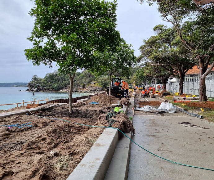 Workmen and saplings in fresh dirt along a promenade under construction, overlooking a small picturesque bay.