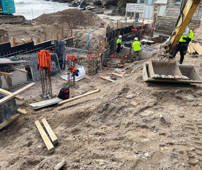 Construction machinery and materials, workers in high-visibility gear and helmets, and a grey frothy sea just visible in the background.