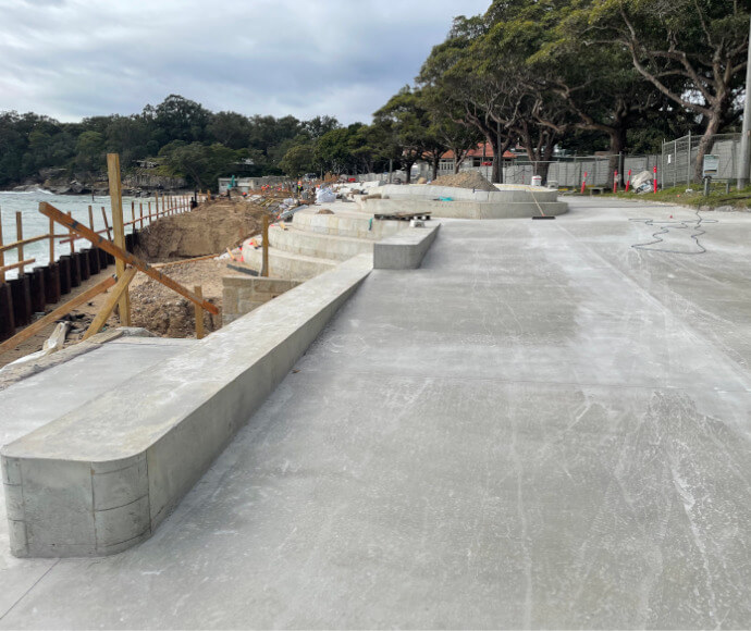 A smooth, freshly paved area of concrete with stepped areas beside a thickly treed shoreline and a grey sky, grey sea in the background.